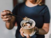 Een persoon in een donkerblauw shirt houdt een beker met dessert of snack vast en schept met een zwarte plastic lepel, mogelijk ijs of yoghurt met toppings.