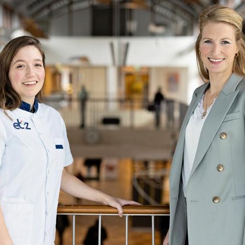 Links een vrouwelijke zorgmedewerker in wit uniform, rechts een vrouw in een lichtgrijze colbert staan aan een ballustrade