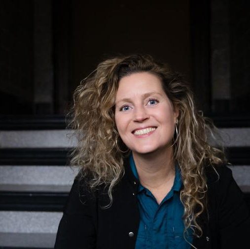 Portret van vrouw met lange, lichtbruine krullen en een zwarte jas met een lichte trap op de achtergrond