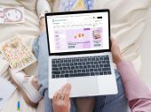 Een vrouw met een laptop op schoot die een webpagina open heeft staan van De Online Drogist. Op de pagina zijn verschillende zonnebrand aanbiedingen te zien.