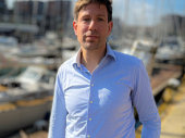 Een man in een blouse staat buiten in de zon, met op de achtergrond zeilboten in een haven.