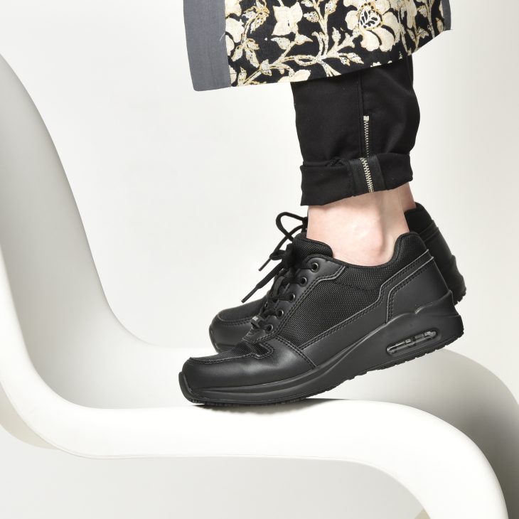 Een persoon staat op een stoel, alleen de schoenen zijn zichtbaar. Het zijn zwarte sneakers en deze persoon staat op zijn tenen.