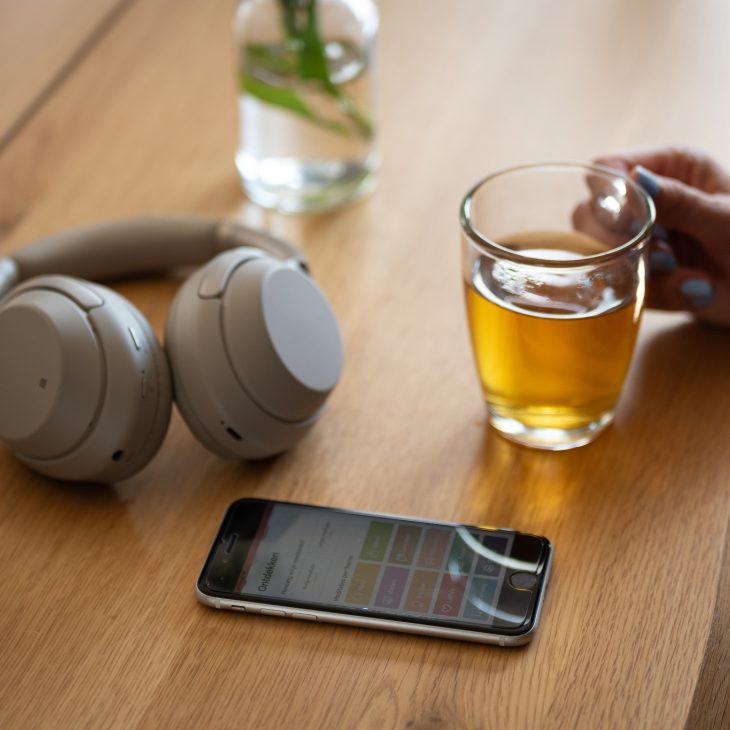 Een grijze koptelefoon ligt op een houten tafel naast een smartphone en een kop dampende thee.