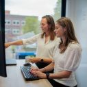 Twee vrouwen met donker haar en witte shirts kijken gezamelijk naar een computerscherm. we zien ze van de linkerkant, op de achtergrond een raam.