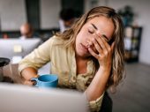 Een vrouw met blond haar zit achter haar computer en houdt vermoeid haar hand voor haar ogen.