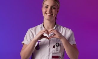 Lachende zorgmedewerker in uniform maakt hartgebaar met handen voor paarse achtergrond.